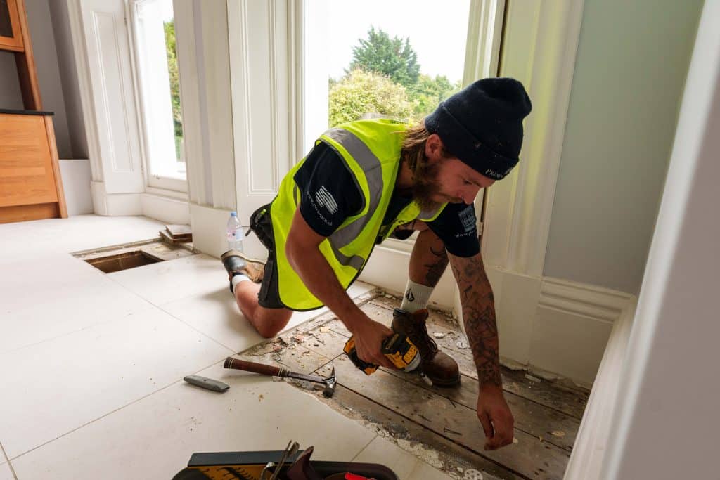 carpenter working on a project, nealing on the floor and using a drill to fix the wooden flooring. wearing a yellow highvis and a blue presence & co. beanie