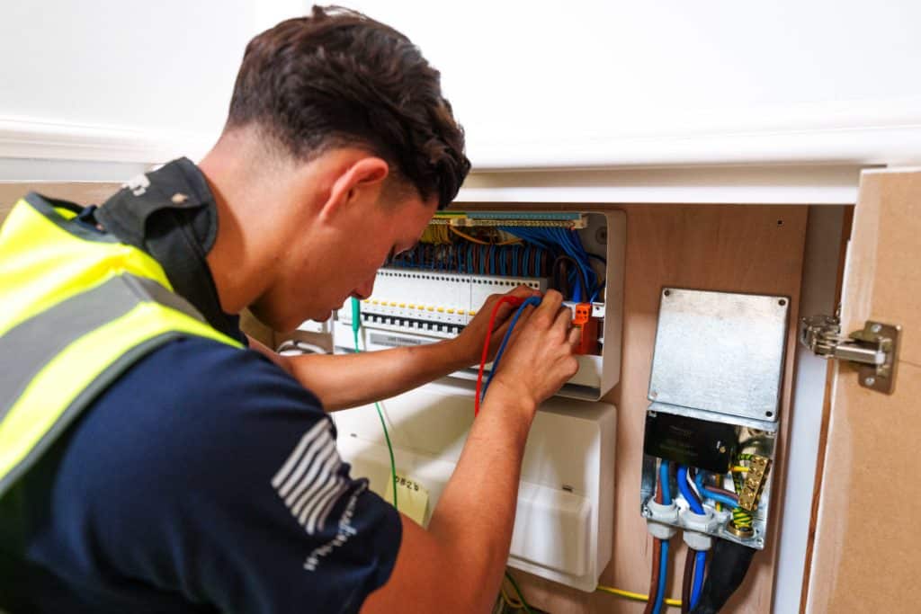 Electrician knelt on the ground wearing a yellow highvis and navy blue Presence & Co. top. Holding a screwdriver., he is testing a consumer board.