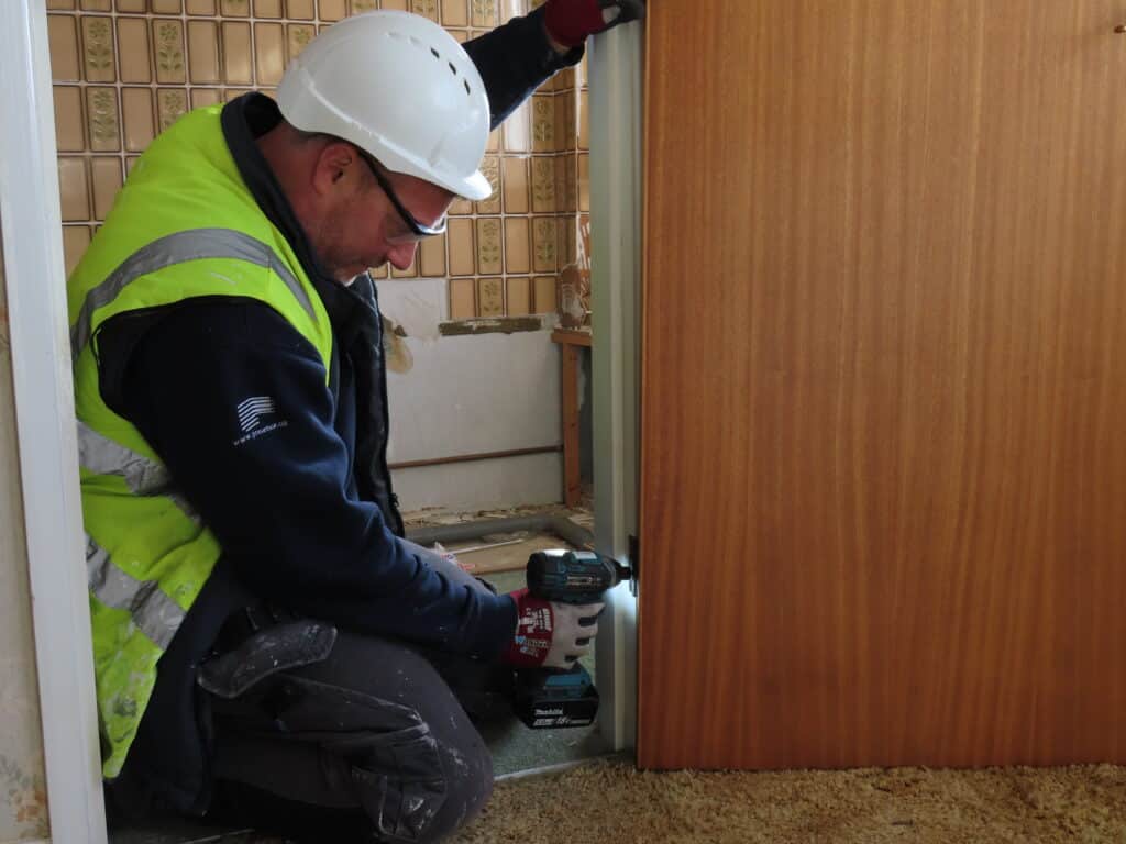 Technician kneeling down on the floor, wearing a green high vis and a white helmet for PPE. Drilling holes into a oak door ready to fix back onto the wall.
