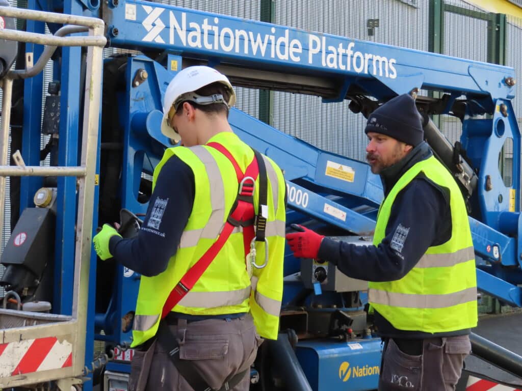 Aaron our electrician in yellow higvis with red straps around his back in PPE with hard hat and gloves. Preparing to get on a blue cherry picker, with other electrician on the ground for safety.