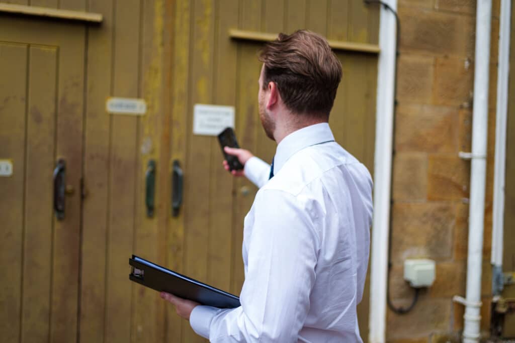 man in a white shirt taking pictures of a wooden building door, holding a black notepad and mobile phone