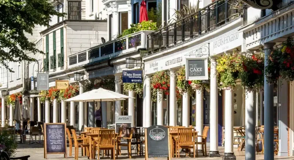 tunbridge wells pantiles in the sun, featuring shops and cafes, surrounded by trees and hanging flowers and tables and chairs outside. written menu boards