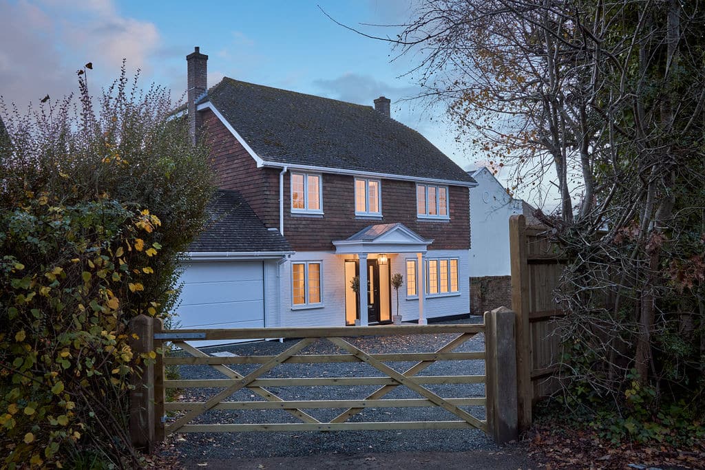 External photo of the house which white on the ground floor and brick on the first floor taken at dusk with downstairs lights on and gate of house in view